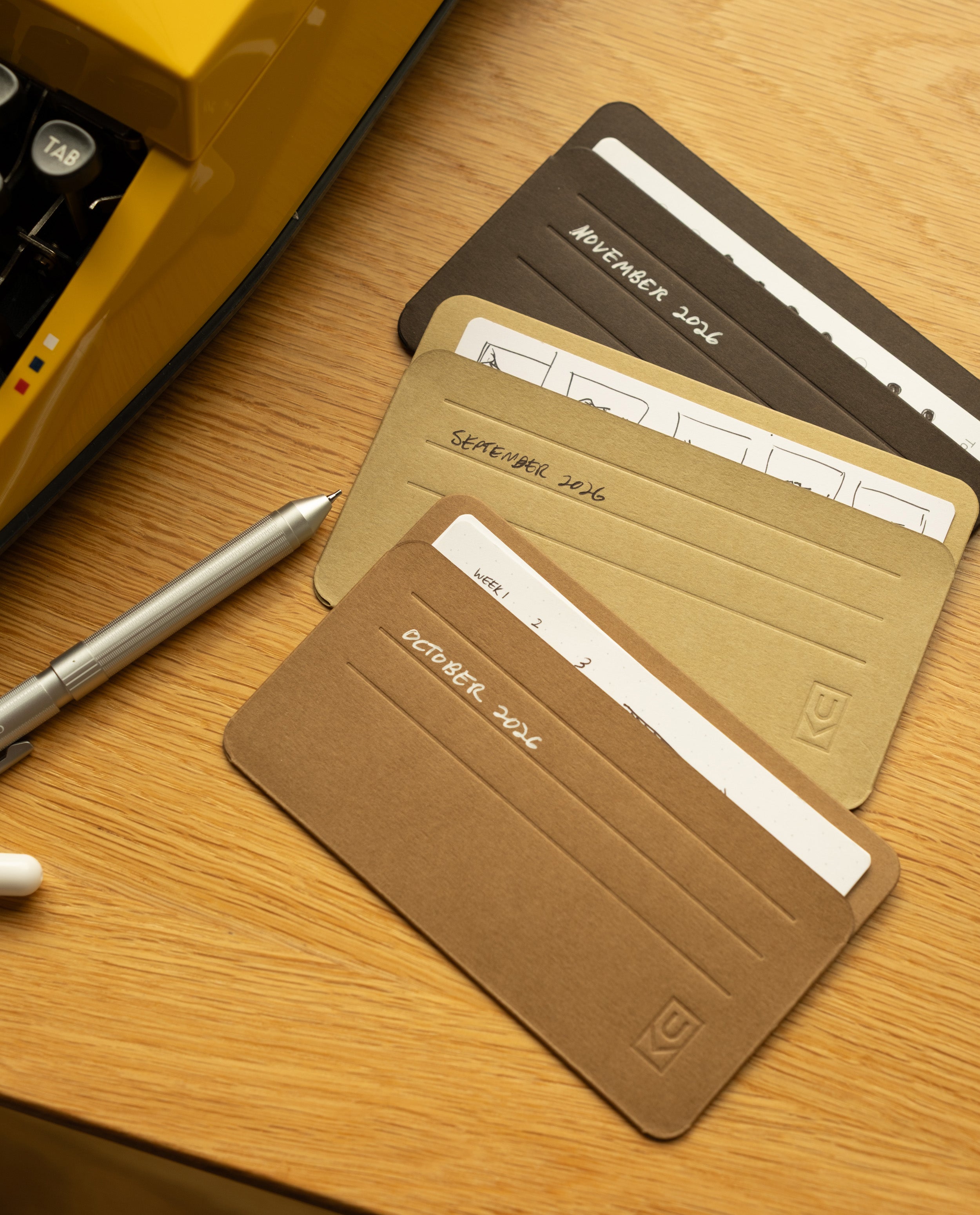 Three labeled monthly planner cards—October, September, and November 2024—are spread on a wooden desk next to a silver pen and a yellow typewriter. Each card fits neatly into your card organizer or Analog Card Sleeves - Small (Earth), with white spaces for notes or schedules.