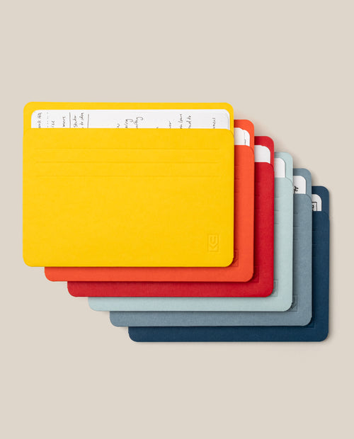 Six brightly colored Large Analog Card Sleeves (Set of 6 - Bright)—in yellow, orange, red, dark gray, light gray, and blue—are stacked together with a handwritten note partially visible in the top yellow sleeve. Ideal for anyone seeking to organize cards efficiently, these vibrant sleeves stand out against a neutral beige background.