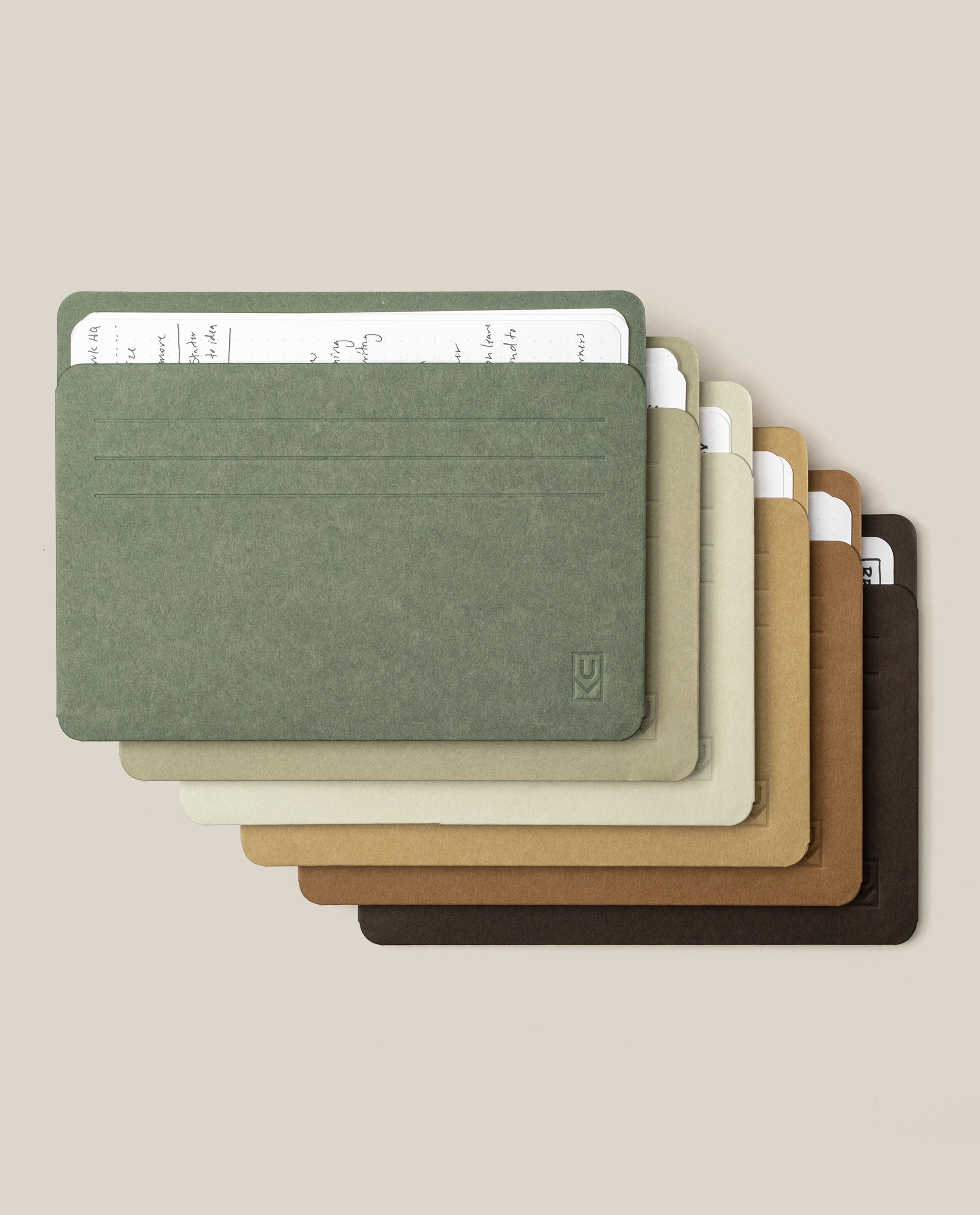 A set of Large Analog Card Sleeves (Set of 6 - Earth tone) is stacked, showcasing minimalist card holders in green, gray, beige, tan, brown, and dark brown. Each sleeve contains a paper card with handwritten notes visible at the top, set against a plain light beige background.