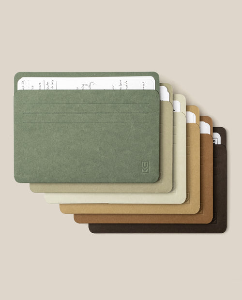 A set of Large Analog Card Sleeves (Set of 6 - Earth tone) is stacked, showcasing minimalist card holders in green, gray, beige, tan, brown, and dark brown. Each sleeve contains a paper card with handwritten notes visible at the top, set against a plain light beige background.