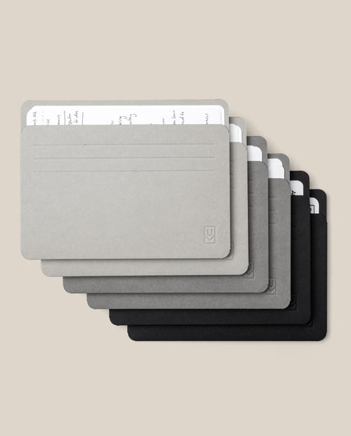 Six Large Analog Card Sleeves (Set of 6 – Monochromatic) in minimalist rectangular shapes and shades of grey and black are stacked in a fan arrangement, each organizer displaying handwritten notes peeking from the top.