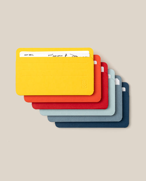 Six Small Analog Card Sleeves (Set of 6 – Bright) in yellow, orange, red, light blue, dark blue, and gray are stacked diagonally on a beige background, with the yellow sleeve holding white cards—offering a stylish and practical solution for card storage or as sleek card sleeves.