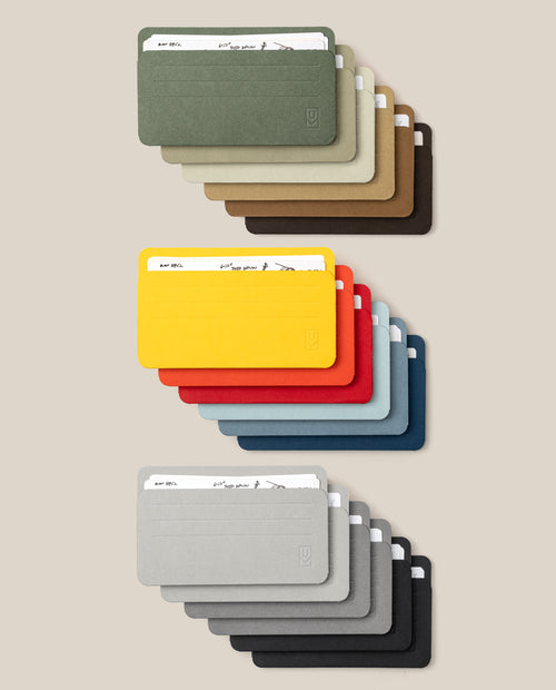 The Small Analog Card Sleeve Multipack (Set of 18) features minimalist card holders and sleeves in a variety of colors, neatly arranged in three rows. Each row displays holders in shades of green, brown, yellow, red, blue, gray, and black against a light beige background—an elegant organizer solution for your cards.