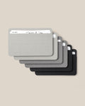 Six Small Analog Card Sleeves (Set of 6 - Monochrome) in shades of gray and black are stacked in a fanned-out arrangement on a beige background, with some cards partially visible inside the sleeves, offering an elegant solution for card organization.
