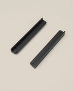 Two black, rectangular Warehouse Item - Capture Tray Clips (Black - Large) with textured surfaces are positioned diagonally apart on a plain light beige background, showcasing their robust design.