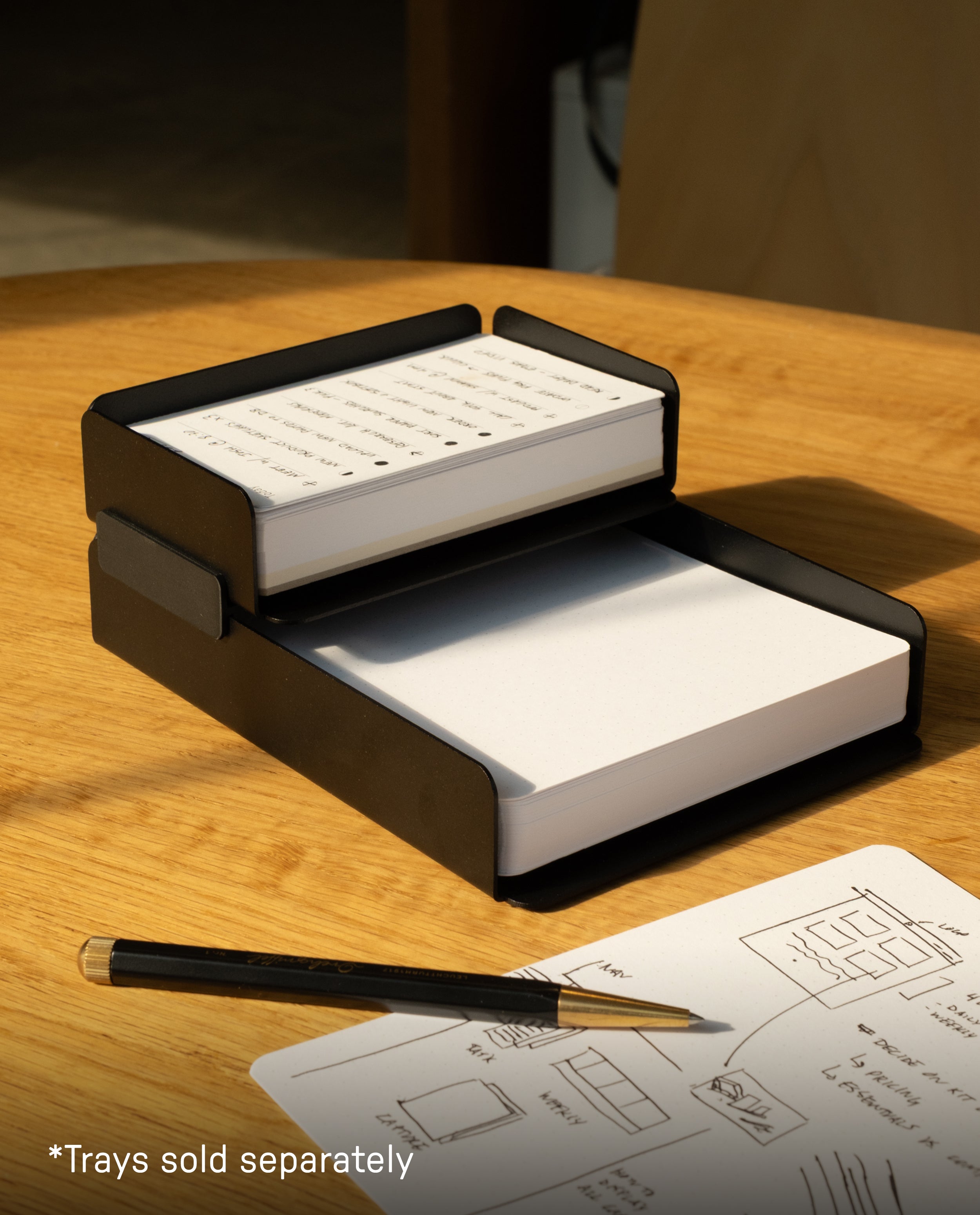 Two black stackable trays hold blank and annotated index cards on a wooden table. A pen and a paper with sketches lie nearby. Text in the corner reads, “*Trays sold separately.”.
