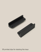 Two black 3D printed clips are displayed on a beige background. Designed for the Capture Bundle (Small Tray + Large Tray - Black Steel), the rectangular clips have raised edges to securely stack the trays. Text: 3D printed clips for stacking the trays.