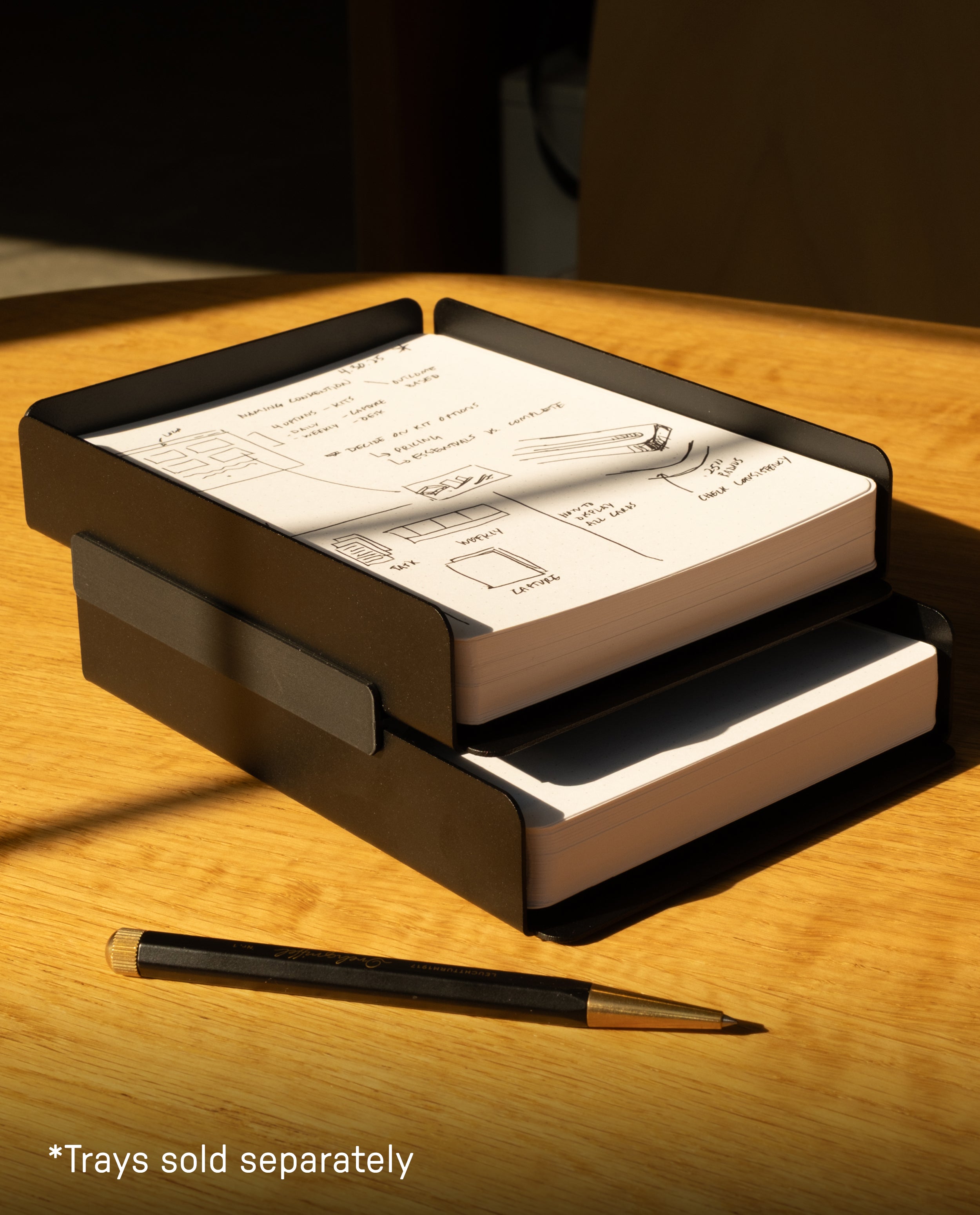 Two black paper trays are stacked on a wooden desk, each containing a notepad filled with sketches and notes. The Warehouse Item - Capture Tray Clips (Black - Large) secure the trays together, providing effortless organization. Nearby rests a black and gold pen. Sunlight streams in, casting shadows across the scene. In the corner, text reads “*Trays sold separately.”.