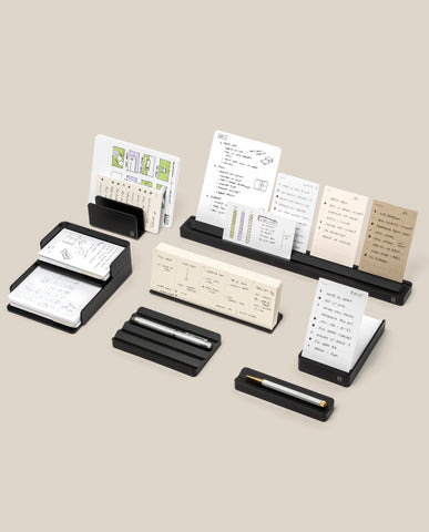 A wooden table displays several minimalist desktop organizers holding note cards with handwritten notes and diagrams, along with a pen and a closed case.