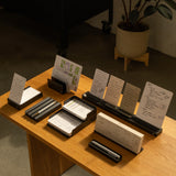 A wooden table displays The Complete Analog Collection (Black Aluminum) desktop organizer, holding note cards, notebooks, pens, and papers with handwritten notes and sketches. A potted plant rests in the background on a concrete floor.