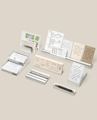 A neatly arranged set from The Complete Analog Collection (Silver Aluminum) displays notecards, papers, and pens on a neutral background, with handwritten notes and schedules in white and beige tones held tidily in minimalist organizers.