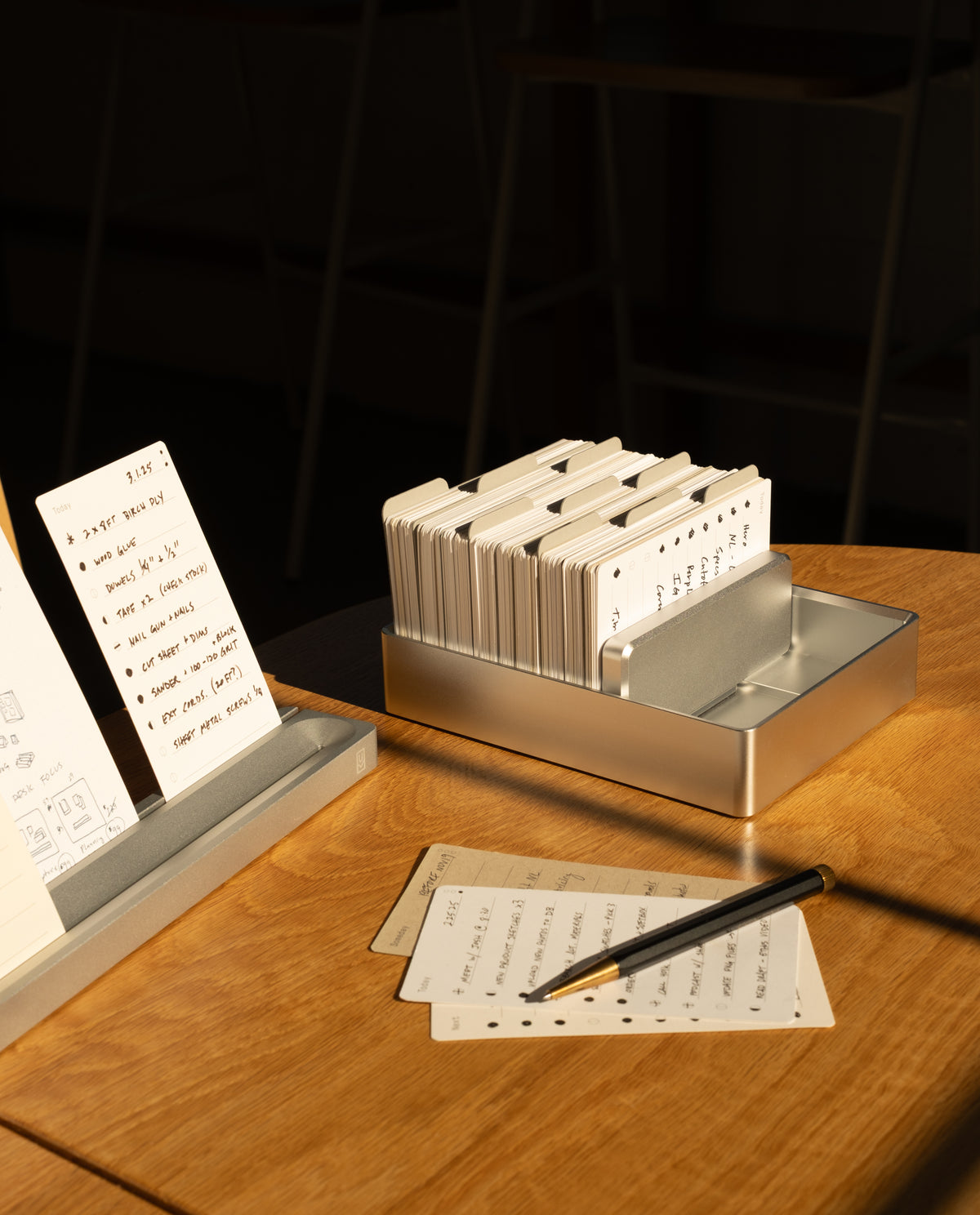 An Analog Archive (Silver Aluminum) filled with index cards, next to a matching stand displaying a handwritten task list and a pen resting on a few loose cards.