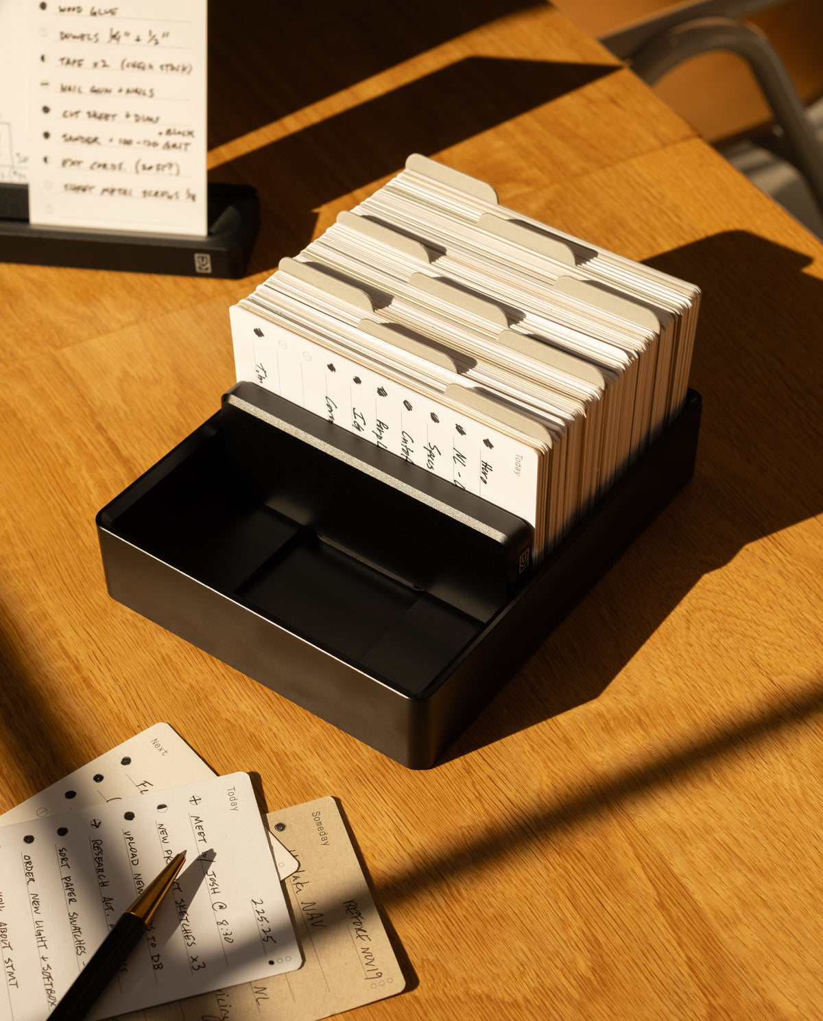 A black Analog Archive (Black Aluminum) card organizer filled with index cards, with a pen and scattered index cards resting on the wooden table surface nearby.