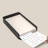 A hand pulls a handwritten card—an Analog Daily Focus Kit + 6 Refills (Black Aluminum)—from a black, rectangular desktop organizer filled with more lined cards against a plain, light beige background.