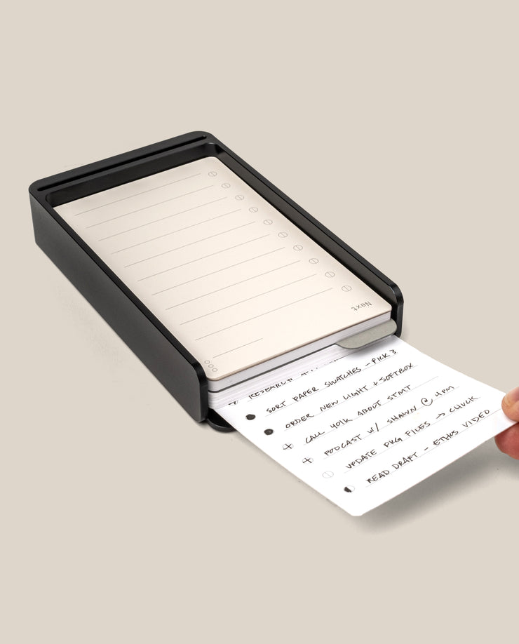 A hand pulls a handwritten card—an Analog Daily Focus Kit + 6 Refills (Black Aluminum)—from a black, rectangular desktop organizer filled with more lined cards against a plain, light beige background.