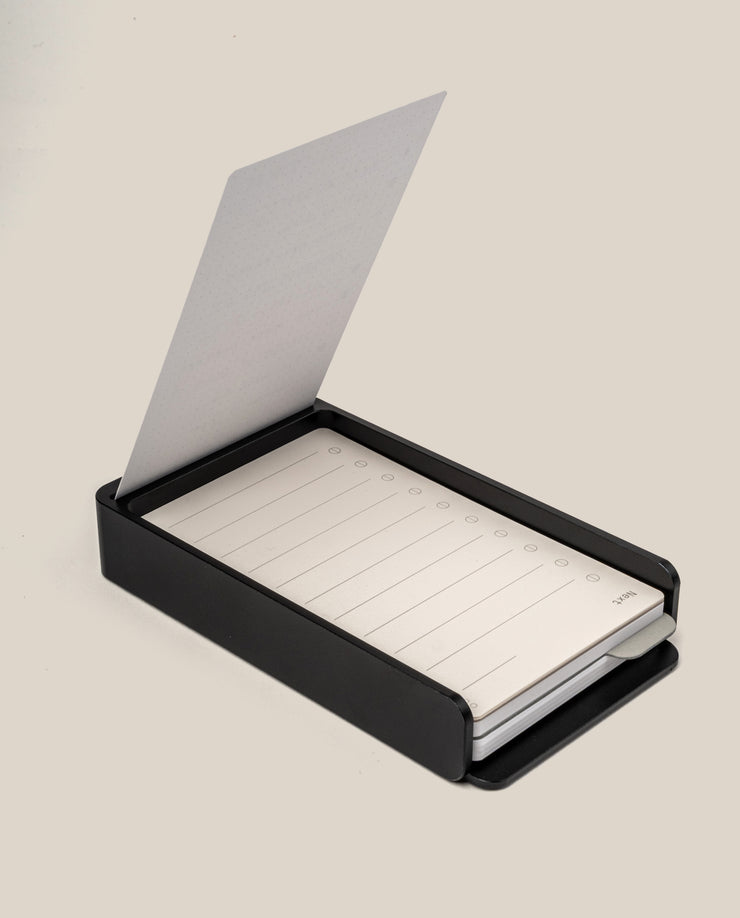 The Analog Daily Focus Kit + 6 Refills (Black Aluminum) features a sleek black rectangular box holding a notepad with lined, numbered sheets; one sheet is flipped open and stands upright.