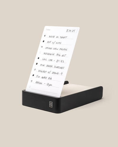 The Analog Daily Focus Kit (Black Aluminum) features a handwritten to-do list card displayed upright in a sleek black aluminum holder on a beige background, helping organize daily tasks as part of a productivity system.