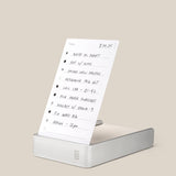 A handwritten to-do list on white paper stands upright in the Analog Daily Focus Kit (Silver Aluminum). Tasks like writing, photo uploads, research, and meetings are neatly penned in black ink. Background is light beige.