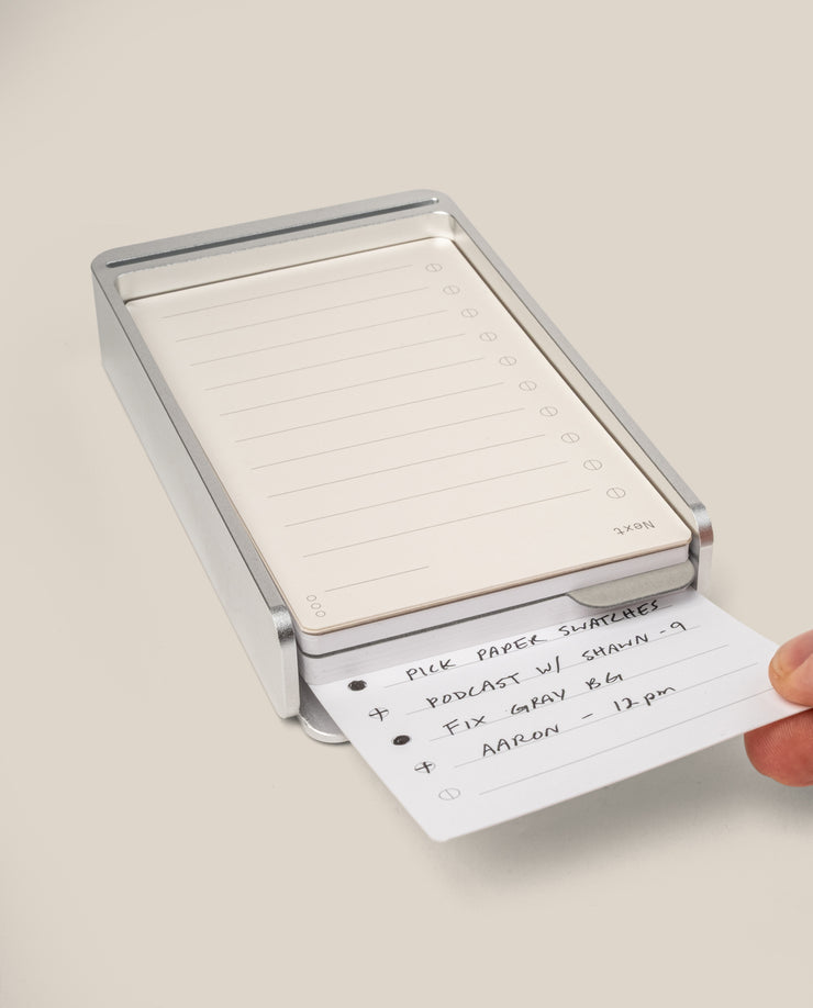 A hand pulls a task card from the Analog Daily Focus Kit + 6 Refills (Silver Aluminum), revealing a handwritten to-do list on a lined card—an elegant analog productivity system against a clean, light-colored background.