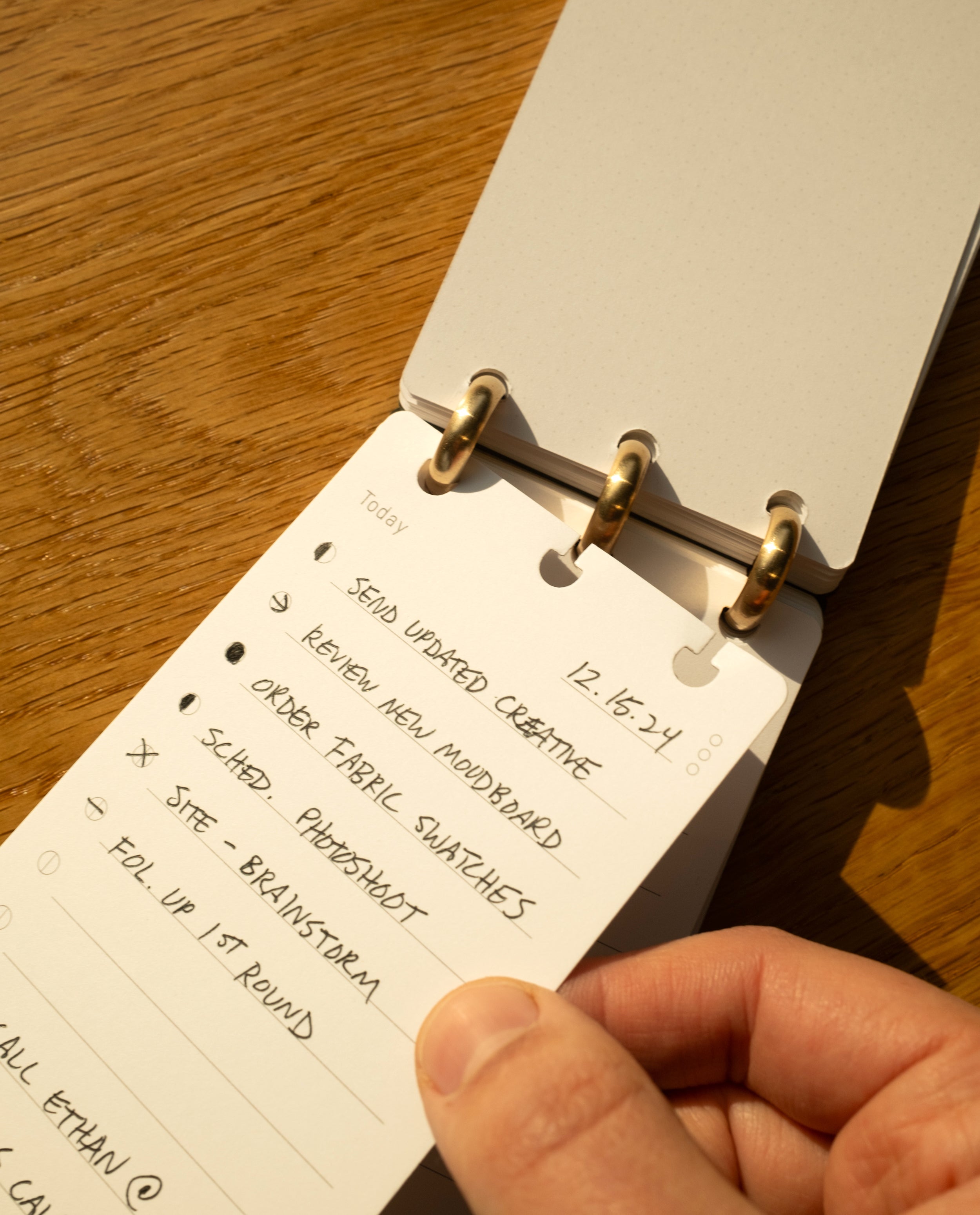 Close-up of a hand holding a Discbound Analog + 3 Refills planner on wood, displaying a handwritten to-do list for 12-16-24 with tasks like creative work, moodboard, fabric swatches, and photoshoot.