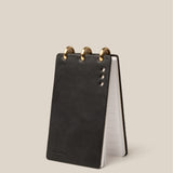 A compact Leather Discbound Analog (Black) notebook with three brass rings on top stands upright on a light beige background. Visible metal rivets and lined pages inside make it ideal for organizing Analog Task Cards.