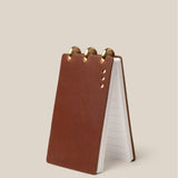 The Leather Discbound Analog (Brown) is a compact flip notebook featuring full-grain leather covers, metal discs at the top, and lined pages, displayed upright on a light beige background.