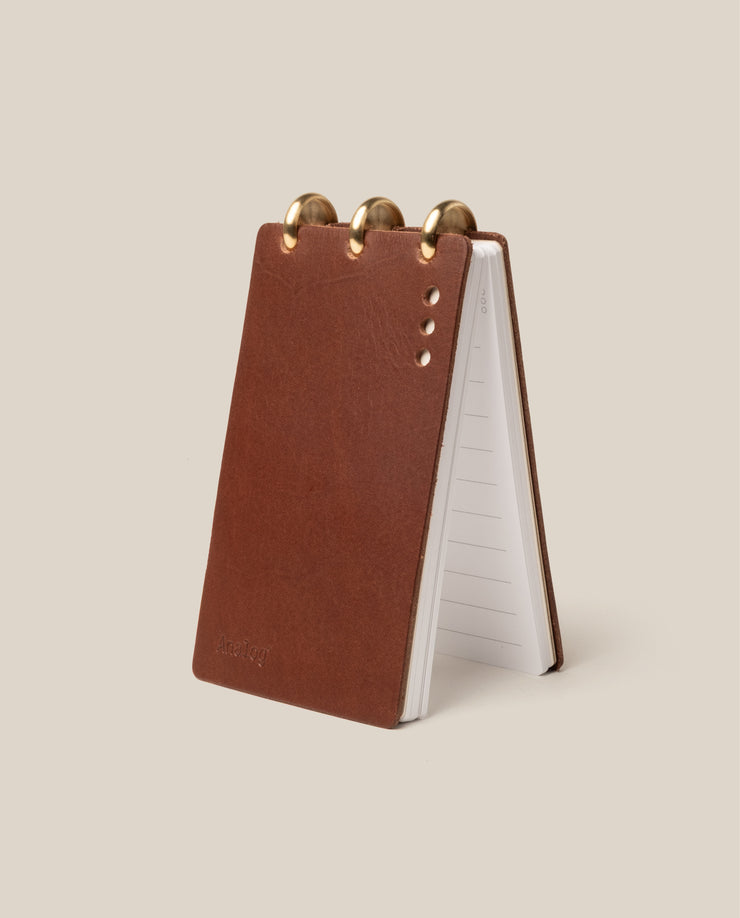 The Leather Discbound Analog (Brown) is a compact flip notebook featuring full-grain leather covers, metal discs at the top, and lined pages, displayed upright on a light beige background.