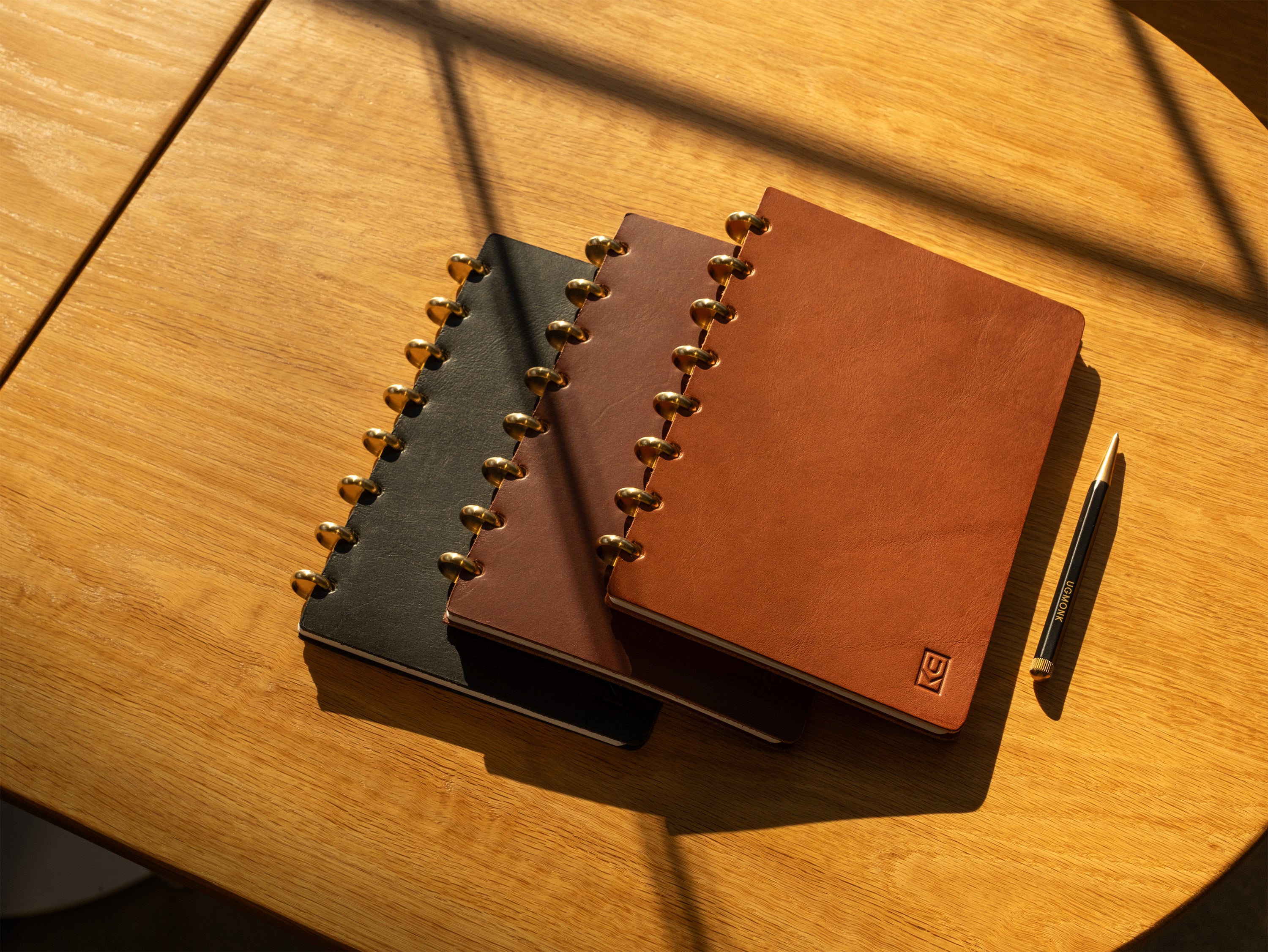 Three leather-bound notebooks, one black and two shades of brown, are stacked on a wooden table next to a pen. Sunlight casts shadows across the scene.