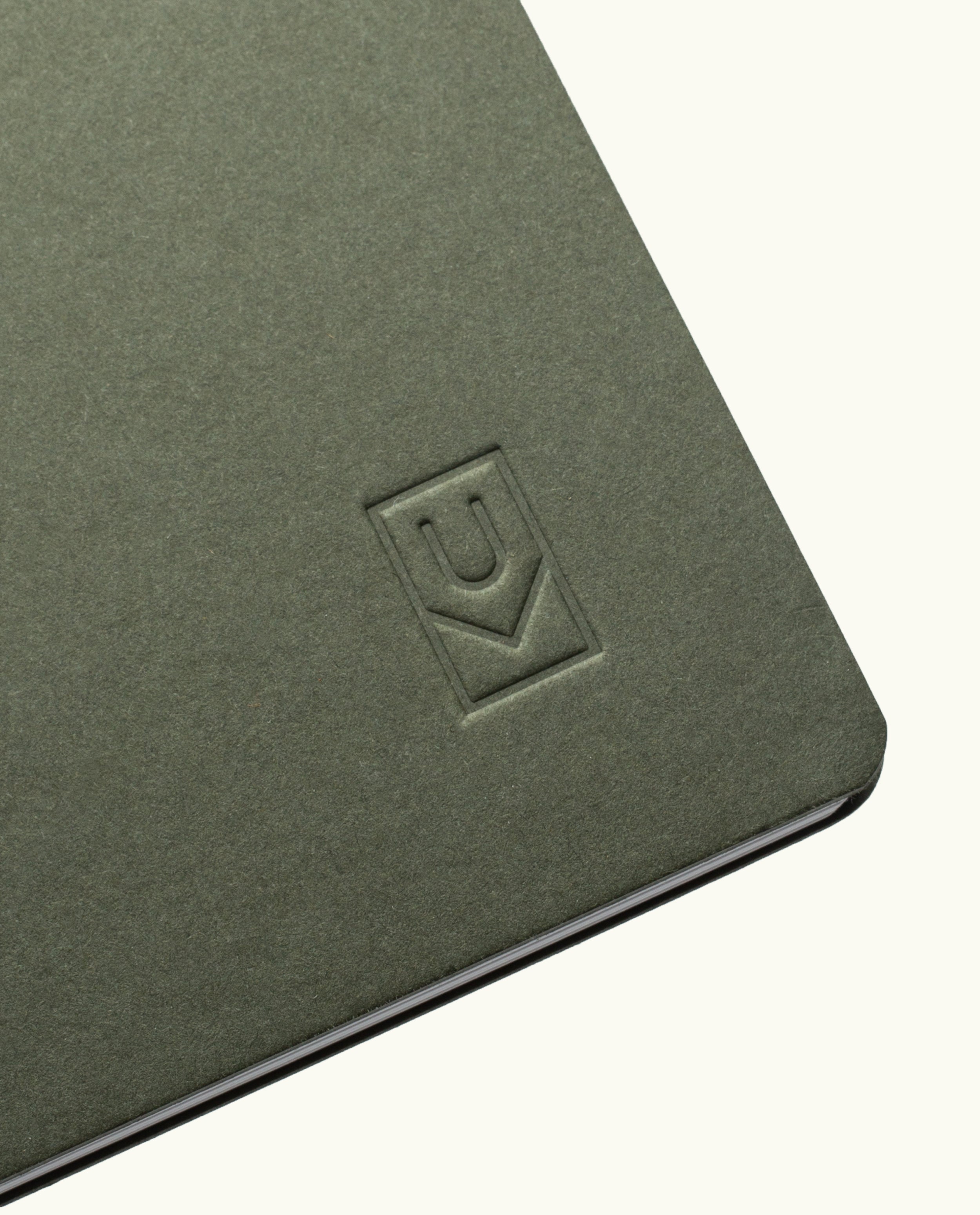 A close-up of the corner of the Discbound Journal (Standard 6"x 8.5") in green, featuring an embossed geometric logo on the cover and solid brass discs binding partially open white pages inside.