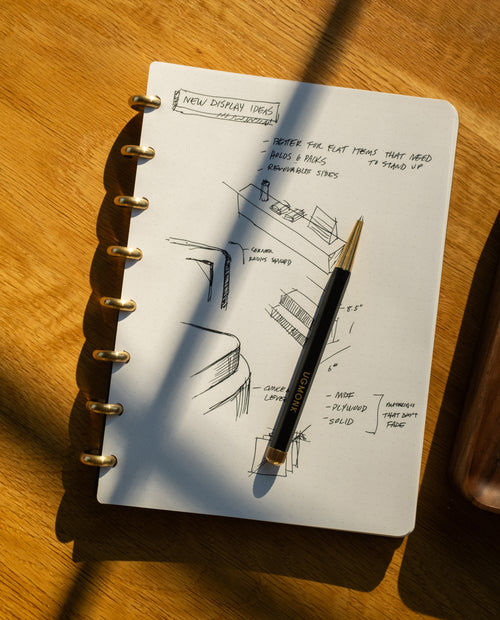 A Discbound Journal + 3 Refills (Standard 6″x 8.5″ Journal) sits on a wooden table, open to dot grid paper filled with sketches and notes titled NEW DISPLAY IDEAS, while a black pen rests on its refillable pages as sunlight casts shadows across the scene.