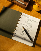 A Discbound Journal (Standard 6"x 8.5") with sketches and notes on architectural design sits open on a wooden desk, a black and gold pen resting on its refillable pages as sunlight casts shifting shadows across the scene.
