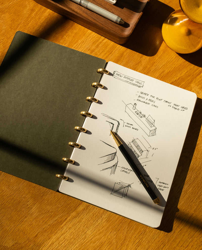 A Discbound Journal + 3 Refills (Standard 6″x 8.5″ Journal) lies open on a wooden desk in warm sunlight, filled with design sketches and notes, with a black and gold pen resting on its page. Nearby, a tray holds assorted stationery items, while the corner of a yellow object is visible.