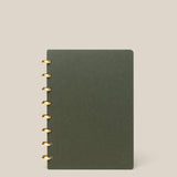 A Discbound Journal (Standard 6"x 8.5") in dark green with brass discs on the left stands upright against a plain beige background.