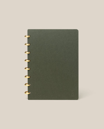 A Discbound Journal (Standard 6"x 8.5") in dark green with brass discs on the left stands upright against a plain beige background.