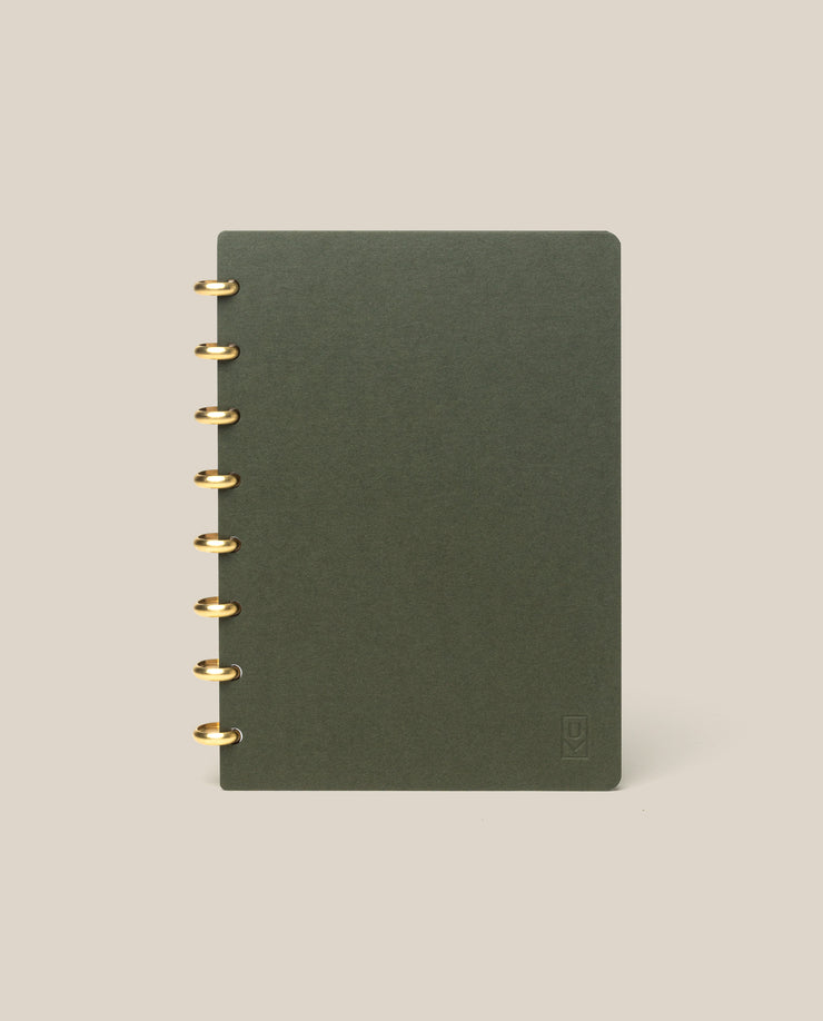 A Discbound Journal (Standard 6"x 8.5") in dark green with brass discs on the left stands upright against a plain beige background.