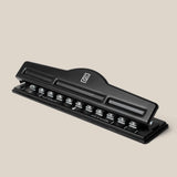 A black metal Discbound Punch with a lever handle sits at an angle on a neutral beige background—ideal for prepping pages for your Discbound Punch or any discbound system.