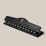A black metal Discbound Punch with a lever handle is displayed on a light beige background. It features measurement markings and metal cutters for punching holes compatible with the Ugmonk Discbound Journal or any discbound system.