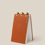 The Leather Discbound Analog (Tan) stands upright on a beige background, featuring a full-grain leather cover with four metal rivets, three brass rings, lined pages, and "Analog" embossed in the bottom corner.