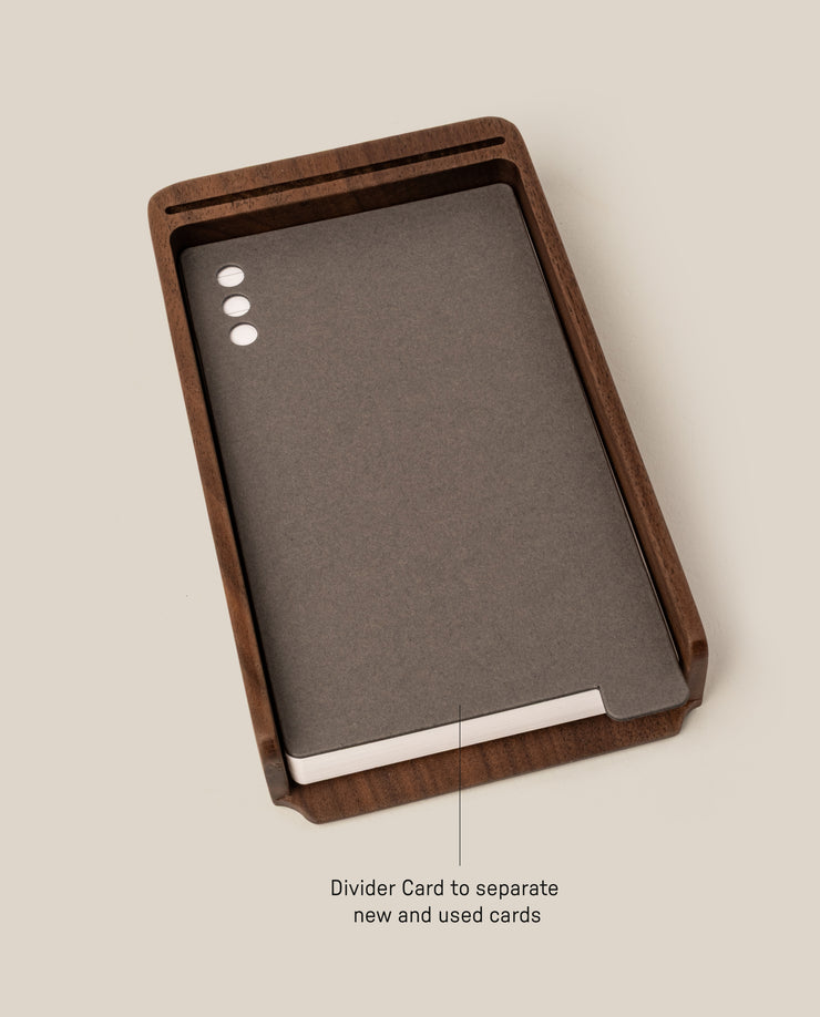 The Analog Daily Focus Kit (Walnut) features a wood card holder displaying dark gray analog task cards with three white dots, organized by a labeled divider for new and used cards, creating a simple productivity system on a light beige background.