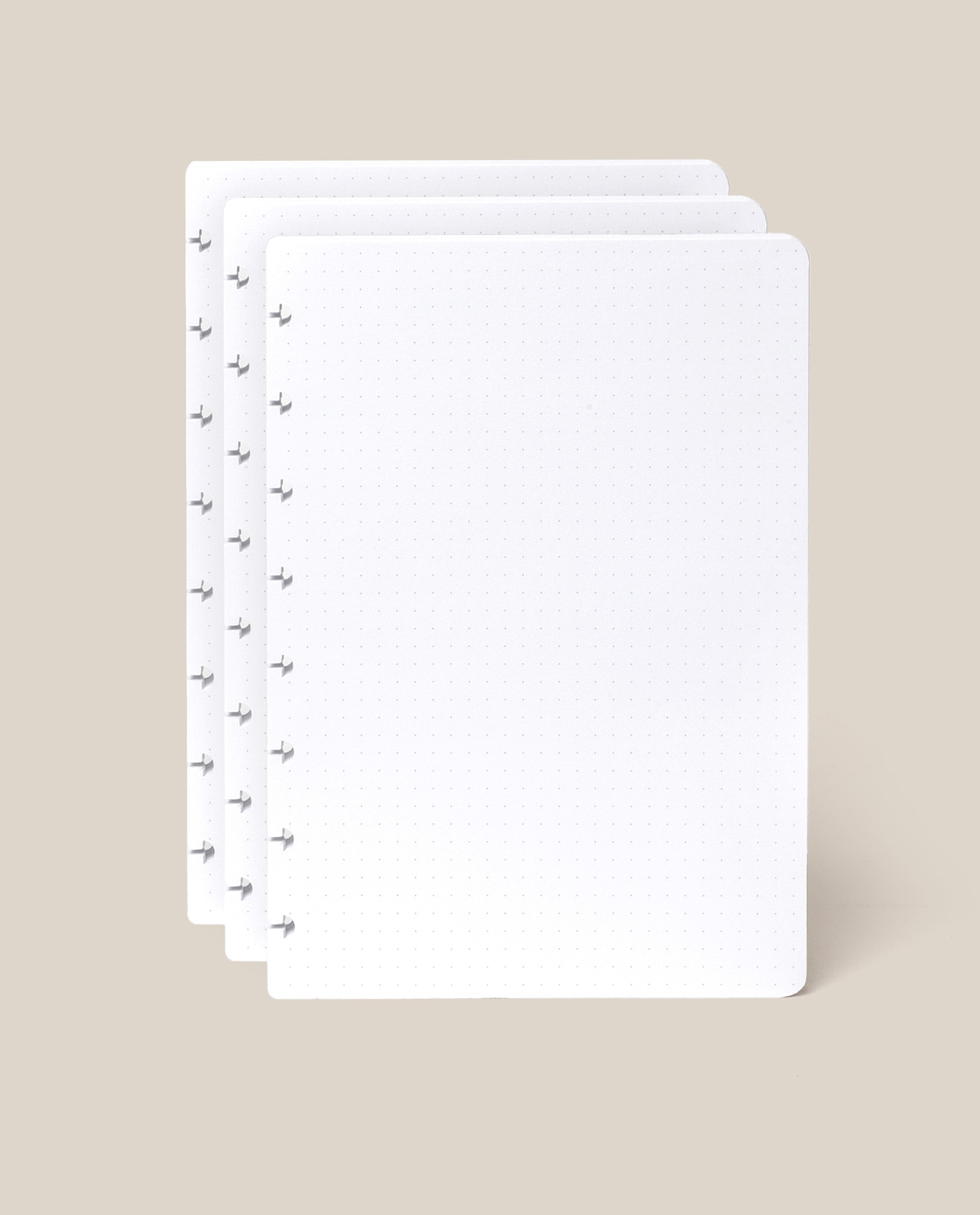 Three sheets of white, dot grid paper with rounded corners are stacked slightly offset on a beige background. The left side of each sheet features several small disc-binding holes, making them perfect as the Discbound Journal Refill - Dot Grid (Standard 6″ x 8.5″) for your Heirloom Discbound Journal or as part of a convenient journal refill pack.