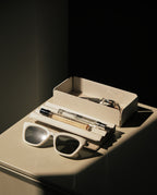 A pair of white sunglasses, a pen, and a small perfume bottle sit on the Gather Medium Tray (2x1 - White) next to an open cream-colored box, all highlighted by dramatic sunlight and shadows.