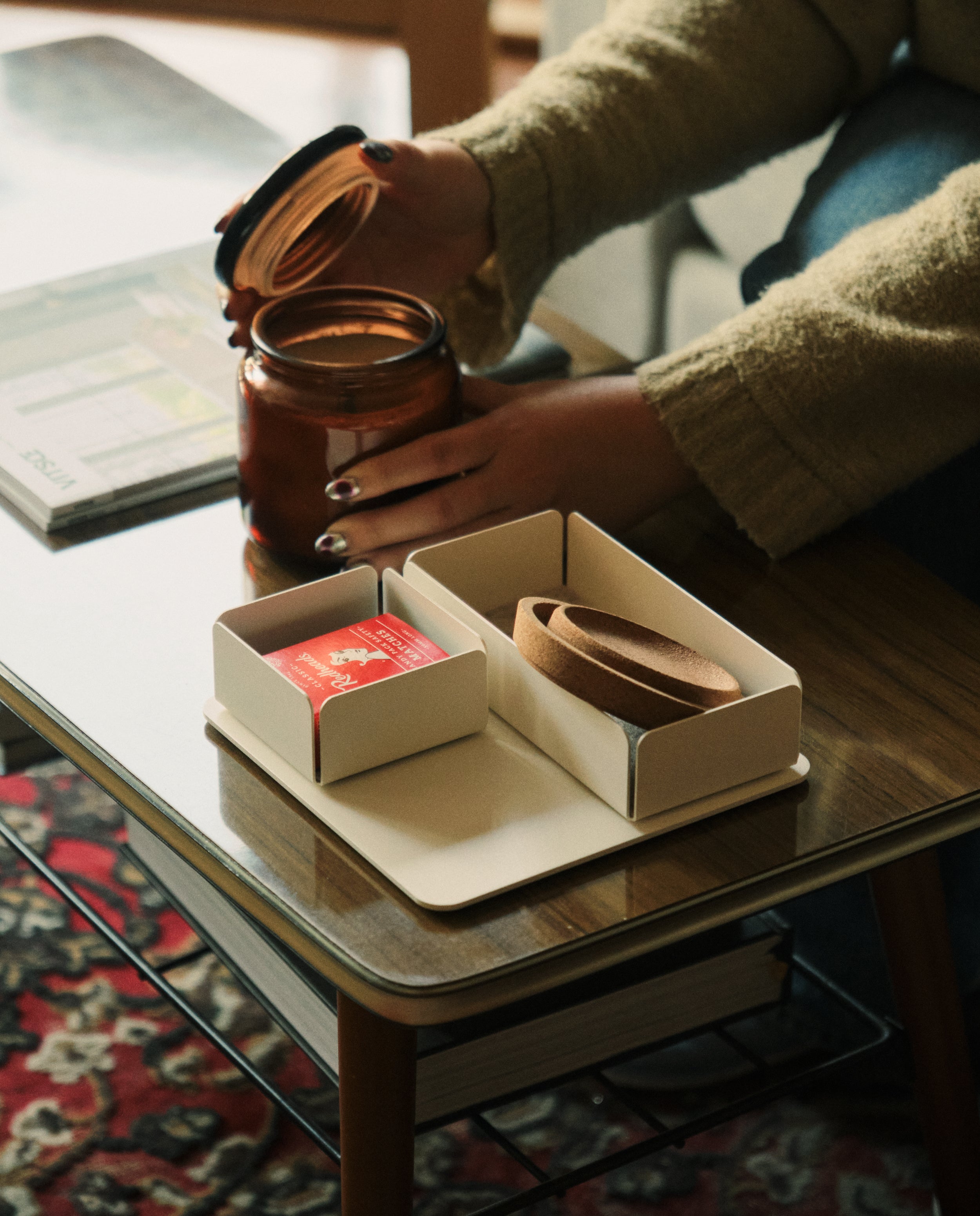A person with dark nail polish opens a glass jar on a wooden coffee table. On the table are two small boxes, including the Gather Medium Tray (2x1 - White), one holding a red matchbox and the other containing a brown leather strap.