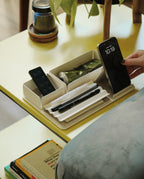 A hand picks up a smartphone from the Gather Medium Tray (2x1 - White), a powder coated steel desk organizer holding pens, a remote, and cream on a yellow table. In the background are a plant, cup, and books on a lower shelf.