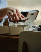 A person with dark nail polish opens a small wooden box beside a brown glass bottle and a white desk organizer. The Gather Small Tray (1x1 - White), made in Pennsylvania, completes the softly blurred background scene.