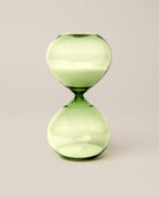 The 15 Minute Hourglass (Green), featuring green glass filled with white sand, stands upright on a simple, light background—serving as an elegant analog timer and a stylish focus tool.