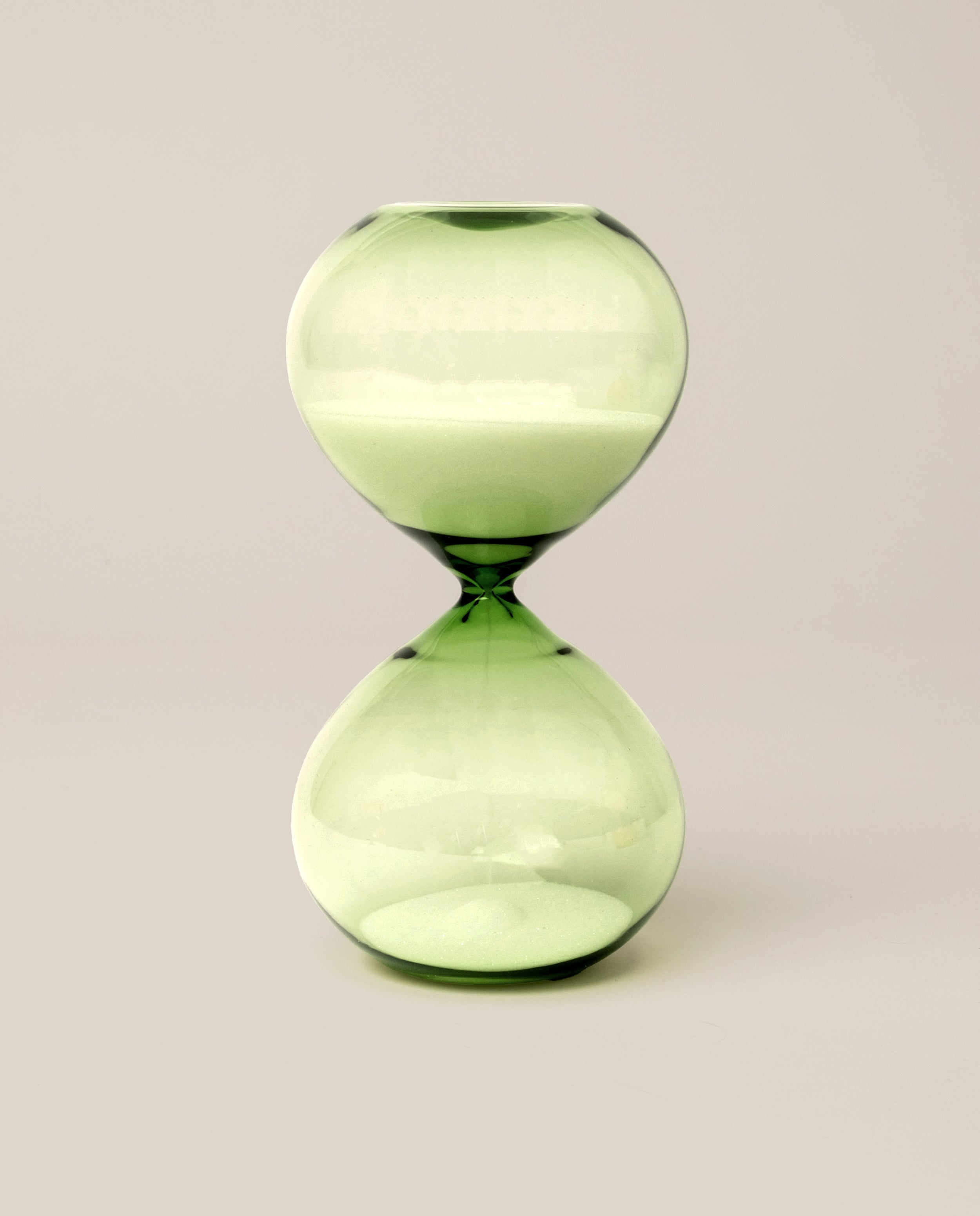 The 15 Minute Hourglass (Green), featuring green glass filled with white sand, stands upright on a simple, light background—serving as an elegant analog timer and a stylish focus tool.