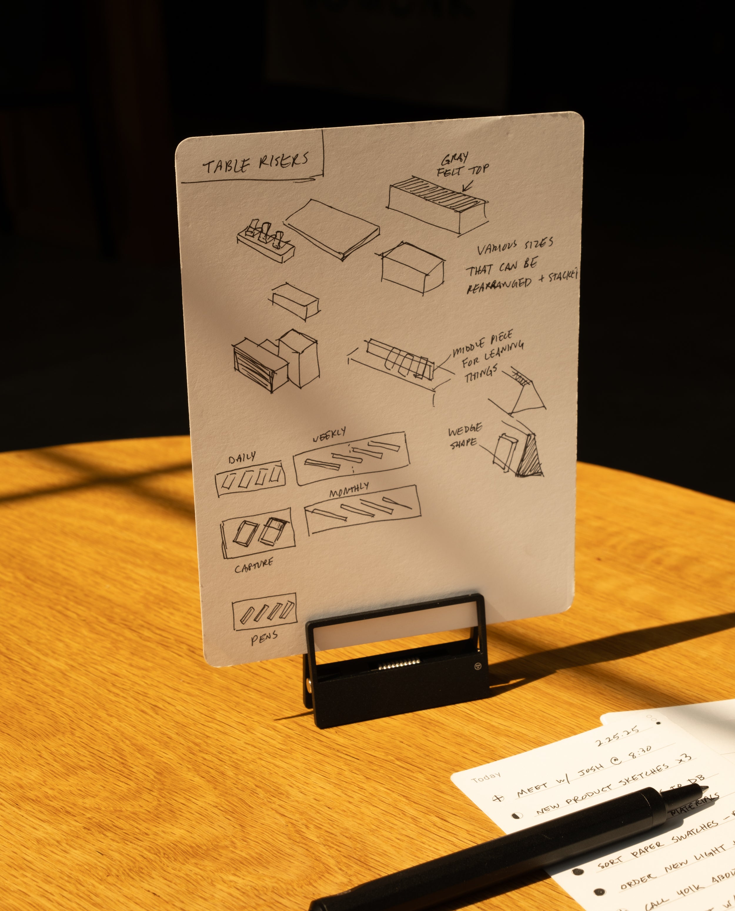 A sheet of paper, secured with HMM Clips (Set of 2 - Black), displays sketches and notes about table risers on a stand. Another sheet with handwritten notes and a black pen rests on a warmly lit wooden table.