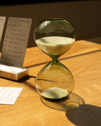 The 15 Minute Hourglass (Green), featuring white sand in a green glass design, sits on a wooden table and casts a shadow, with blurred handwritten notes and a menu visible in the background.