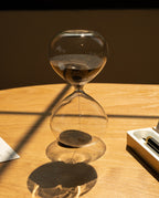 The 15 Minute Hourglass (Clear), filled with black sand, rests on a wooden table and casts a distinct shadow in the sunlight. Most of the sand remains in the top section, with some already settled at the bottom.