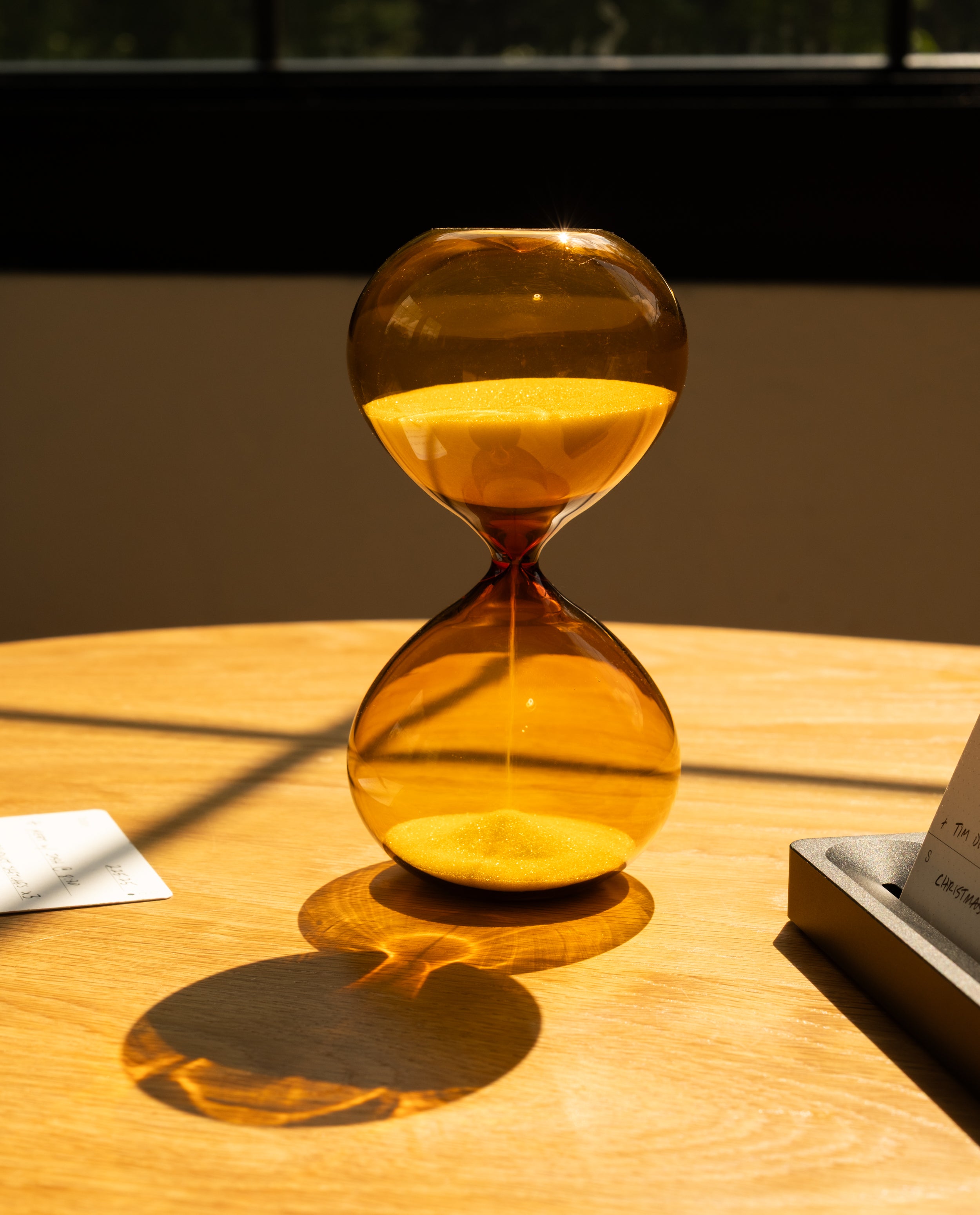 A 15 Minute Hourglass (Amber), designed as a distinctive focus tool, rests on a wooden table with amber sand flowing consistently and casting a defined shadow in the sunlight. Papers and a tray are partially visible nearby, while a window frames the background.